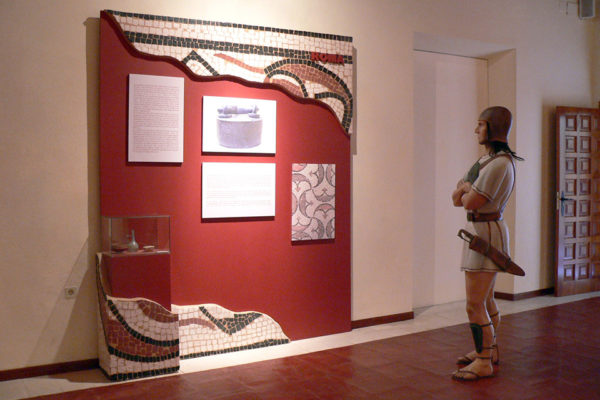 Figura de guerrero íbero contemplando la panelería de estilo romano (rojo pompeyano y mosaico).