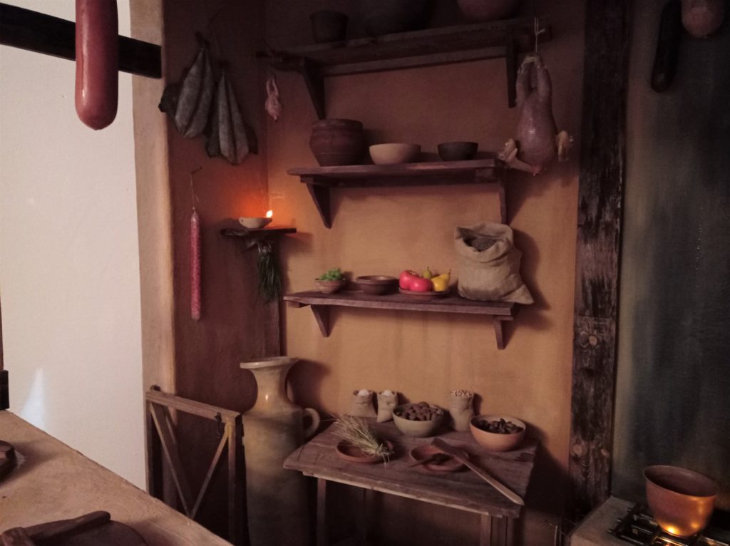 Mesa y estantes con diversos alimentos y ánfora.