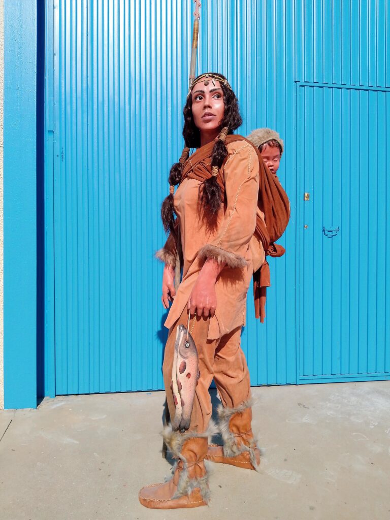 Mujer vestida llevando el bebé en la espalda, con truchas pescadas y arpón