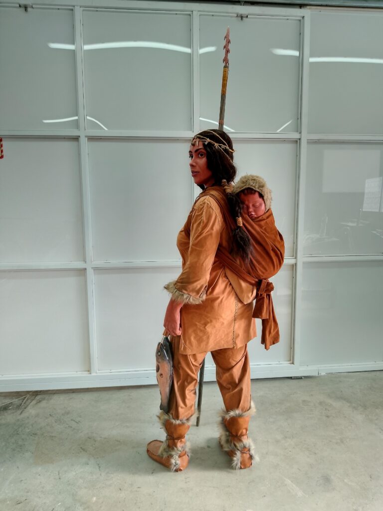 Mujer vestida llevando el bebé en la espalda, con truchas pescadas y arpón